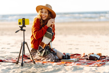 Wall Mural - Young woman with smartphone on tripod on sea beach recording video. Happy woman creates content. Travel, blogging, technology concept.