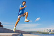 © muse studio - Man trainer in mid-air, leaping on steps with determination, framed by a clear blue sky and urban skyline.