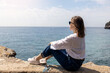 © Margo_Alexa - Young woman in white blouse and jeans, wearing sunglasses, sitting on rock by the sea, looking away on sunny day. Copy space.