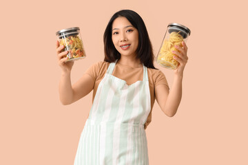 Poster - Happy beautiful Asian woman holding jars of raw pasta on beige background