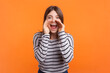 © khosrork - Portrait of positive optimistic happy woman with brown hair keeps hands near mouth screaming making announcement, wearing striped shirt. Indoor studio shot isolated on orange background