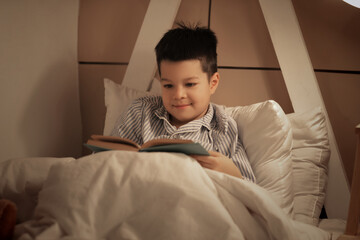 Sticker - Cute little Asian boy in pajamas reading book in bed