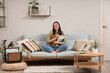 © Pixel-Shot - Young woman reading magazine on grey sofa near light wall at home