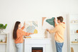 © Pixel-Shot - Young couple hanging paintings on wall above fireplace at home, back view