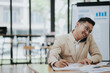 © NanTua - Cheerful young Asian businessman smiling in bright modern office working on laptop computer checking charts, graphs and paper.