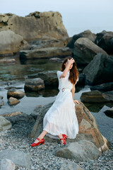 Poster - Serene woman in white dress sitting on seaside rock with vibrant red shoes by ocean view