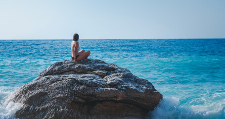 Wall Mural - A young woman in a bikini sits on a large stone on the beach and looks at the sea, relaxing and meditating with nature.