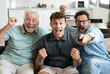© luciano - Enthusiastic three generation men family fans watch football championship or olympic games on TV sitting together on sofa, happy grandson with father and old grandfather celebrate team victory