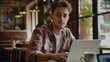 © BigHeartHouston - A young man in a plaid shirt works on his laptop in a cozy cafe. He looks into the frame thoughtfully, surrounded by a warm, inviting interior with wooden furniture, plants, and soft lighting.