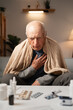 © Andrii Lysenko - Elderly man feeling unwell with pneumonia, wrapped in blanket, holding his chest. Surrounded by medications. Concept of treating pneumonia, cough