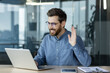 © Tetiana - Smiling young man in blue shirt and glasses sitting at office desk and talking on video call, greeting and waving at laptop camera