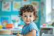 © Irina B - A young child with curly hair holding a crayon, sitting at a table in a colorful classroom with drawings on the wall, looking curiously at the camera..