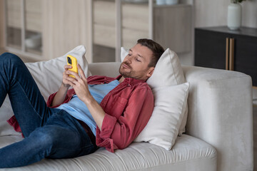 Disinterested man with phone lying on couch surfing online web, procrastination at home. Lazy male with social media addiction on smartphone scrolling internet, enjoy virtual life rest look at screen.