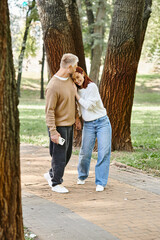 A man and woman in casual attire stand together in a park, surrounded by nature.