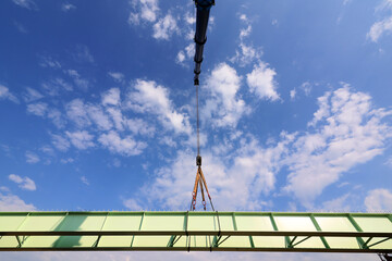 Large tonnage crane lifts a metal section of a bridge under construction against blue sky with white clouds.
