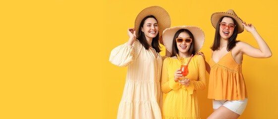 Wall Mural - Beautiful young women in beach hats and with glass of cocktail on yellow background with space for text