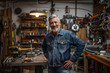 © forenna - The photo captures a middle-aged car mechanic with gray hair and a beard standing in his garage, wearing a jean jacket. With a proud smile, he stands with his hands on his hips. Th