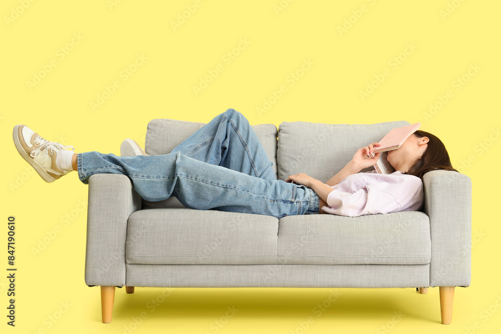 Beautiful young woman with book sleeping on sofa against yellow background