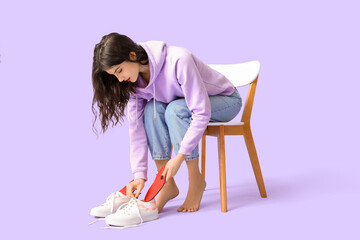 Young woman sitting on chair and putting orthopedic insoles in sneakers on lilac background