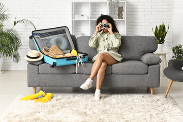 Wall Mural - Beautiful young woman with camera unpacking summer clothes from suitcase at home