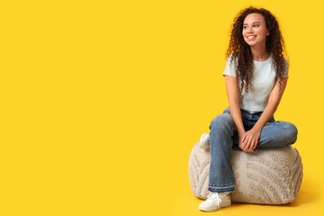 Sticker - Beautiful young happy African-American woman resting on pouf against yellow background