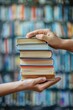 © Naret - Close-up of hands passing a stack of books against a blurred bookshelf background, symbolizing knowledge sharing and book exchange.