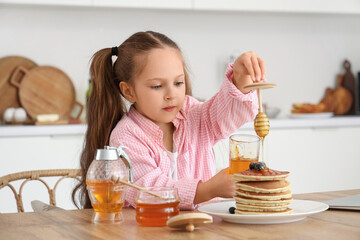 Poster - Cute little girl pouring honey on pancakes at table in kitchen