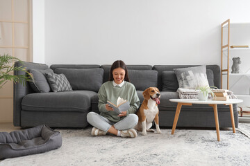 Wall Mural - Young woman reading book and sitting with cute Beagle dog on floor in living room
