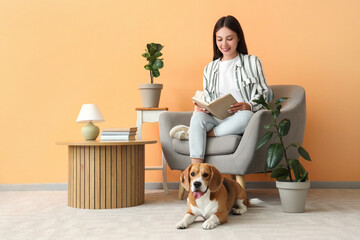 Sticker - Young woman reading book on armchair near adorable Beagle dog resting on floor in living room