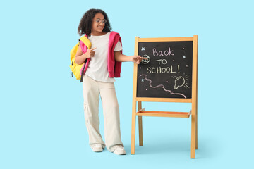 Wall Mural - Little African-American girl pointing at chalkboard with text BACK TO SCHOOL on blue background