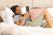 © Wavebreak Media - Biracial young woman relaxing on bed, smiling while using smartphone, at home