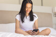 © Wavebreak Media - Biracial young woman sitting on bed, smiling while looking at smartphone, at home