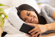 © Wavebreak Media - Biracial young woman lying on bed smiling while looking at smartphone, at home
