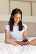 © Wavebreak Media - Biracial young woman sitting on bed, smiling at smartphone, at home