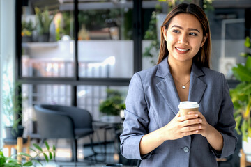 Young happy Asian businesswoman in a suit holding a cup of delivery coffee, standing confident in the office room background