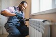 © Serhii - Man in workwear overalls using tools while installing or repairing heating radiator in room