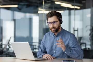 Wall Mural - A serious and focused young man businessman conducts a business meeting via video call, sits in the office in a headset in front of a laptop and explains, gesturing with his hands