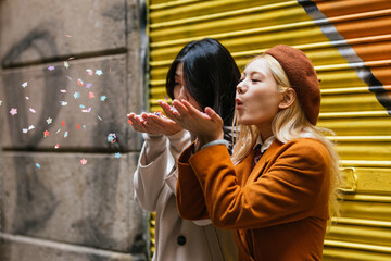 Joyful lesbian couple blowing confetti outdoors on the street.