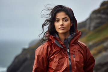 Wall Mural - Portrait of a satisfied indian woman in her 20s wearing a functional windbreaker over dramatic coastal cliff background