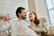 © LIGHTFIELD STUDIOS - A beautiful adult couple, a redhead woman and a bearded man, sit together on a modern sofa in their apartment, enjoying quality time.