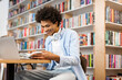 © Home-stock - Positive Black male student reading book in university library sitting at desk with laptop, searching information for project