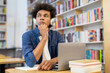 © Home-stock - Pensive African American male student in headphones looking away, creating essay or working on online project sitting with laptop and books in university library