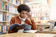 © Home-stock - Concentrated thoughtful latin student guy, sitting at table in campus library, thinking about project or homework, looking at copybook