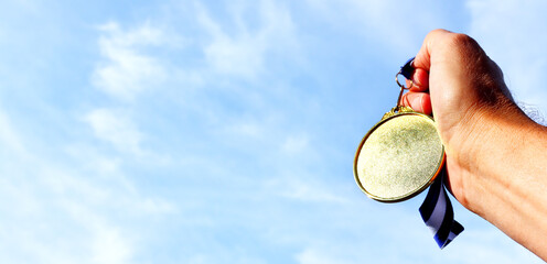 Wall Mural - man hand raised, holding empty gold medal against sky. award and victory concept