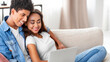 © Prostock-studio - African American teen guy and girl are relaxing on a couch in a living room. The guy is sitting with his arm around the girl, who is looking at a laptop in her lap. They appear happy and comfortable.