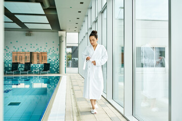 Wall Mural - A young, beautiful brunette woman in a bathrobe standing beside an indoor swimming pool.