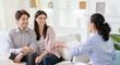 © Prostock-studio - A young couple sits on a white couch, attentively listening to an advisor who gestures while speaking, setting is bright, modern living room with white shelves adorned with minimalistic decorations.