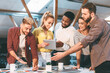 © Prostock-studio - Five colleagues gather around a table in an office, discussing and collaborating on a project using documents and a tablet.