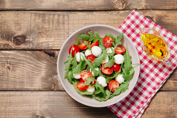 Fresh summer vegetable salad with mozzarella cheese, green arugula leaves and cherry tomatoes on wooden table background. Healthy food, diet concept. Flat lay, top view, copy space.