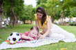 © Ekkasit A Siam - Same park scene, woman and toddler focused on coloring activity, soccer ball and paint supplies on picnic blanket.